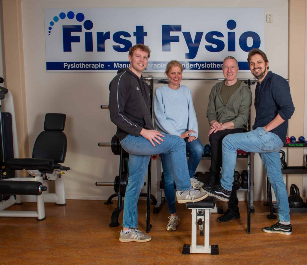 Vier mensen, twee vrouwen en twee mannen, poseren en glimlachen samen in een fysiotherapiekliniek. Ze staan of zitten voor fitnessapparatuur, onder een bord met de tekst First Fysio.