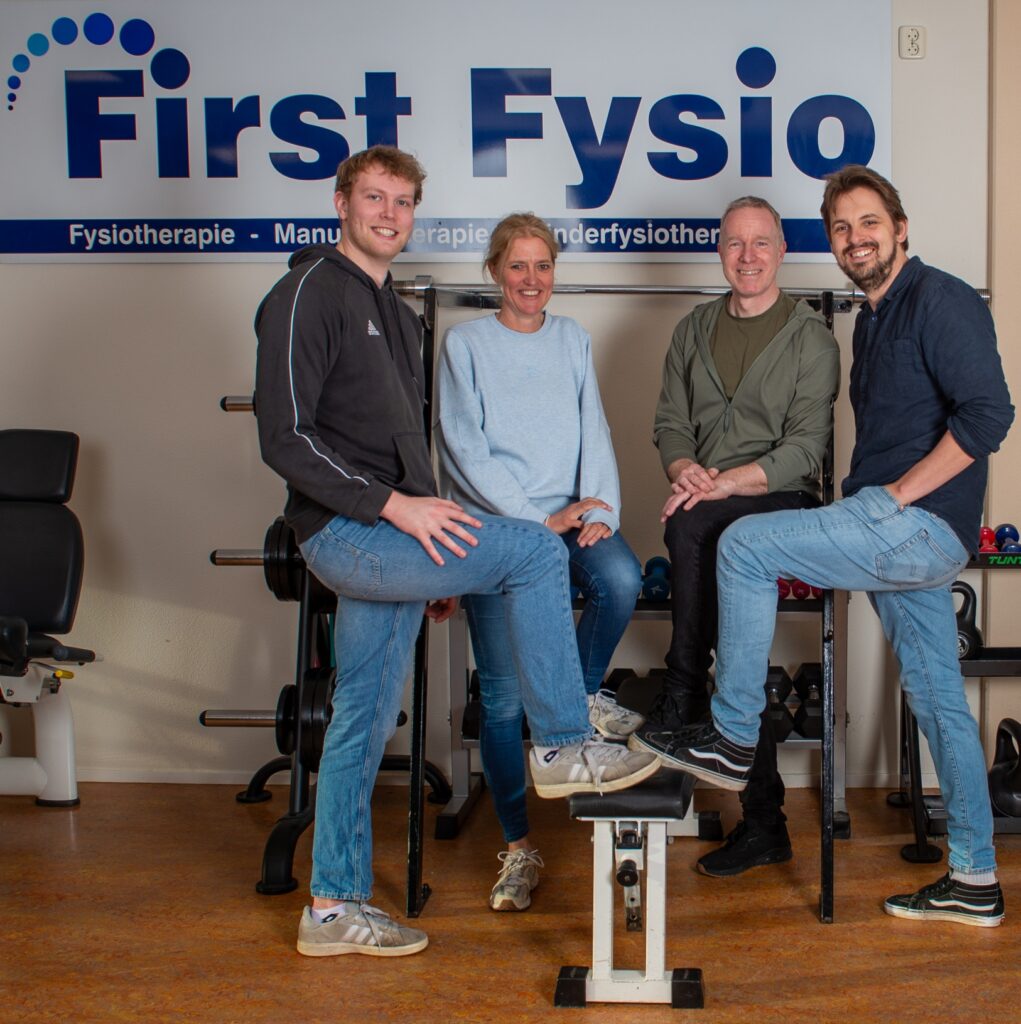 Vier mensen glimlachen en poseren samen in een fysiotherapiekliniek, staand en zittend voor een First Fysio-bord, met fitnessapparatuur en gewichten op de achtergrond.