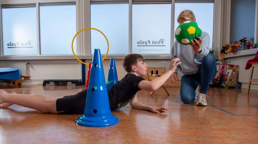 Een kind ligt op de grond en reikt naar een groene en gele bal die wordt vastgehouden door een volwassene in een kamer met blauwe kegels, ringen en ander speelgoed, waarschijnlijk tijdens een fysiotherapie- of speelsessie.