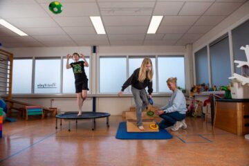 Drie mensen bevinden zich in een helder verlichte kamer. Een jongen springt op een trampoline, een meisje balanceert op een schuimblok terwijl ze een ring vasthoudt en een vrouw knielt vlakbij om het meisje te helpen. Speelgoed en apparatuur zijn zichtbaar op de achtergrond.