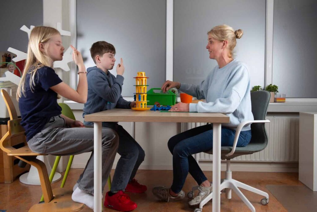 Een vrouw en twee kinderen zitten aan een tafel en spelen samen een bordspel. De kinderen lijken na te denken terwijl de vrouw glimlacht, en een kleurrijk spel met gestapelde stukken ligt in het midden van de tafel.