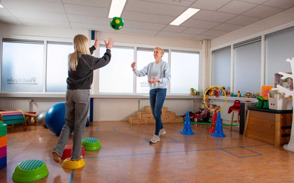 Twee mensen in een helder verlichte kamer spelen vangbal met een groene en gele bal. De een staat op een opstapje terwijl de ander de bal gooit; kleurrijk speelgoed en apparatuur vullen de achtergrond.