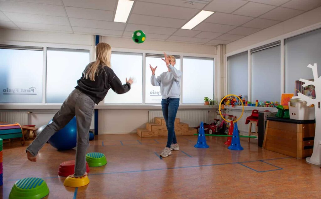 Twee kinderen spelen vangbal met een groene bal in een helder verlichte therapie- of speelkamer, omringd door speelgoed, kleurrijke kegels en balanceerstenen, met grote ramen op de achtergrond.