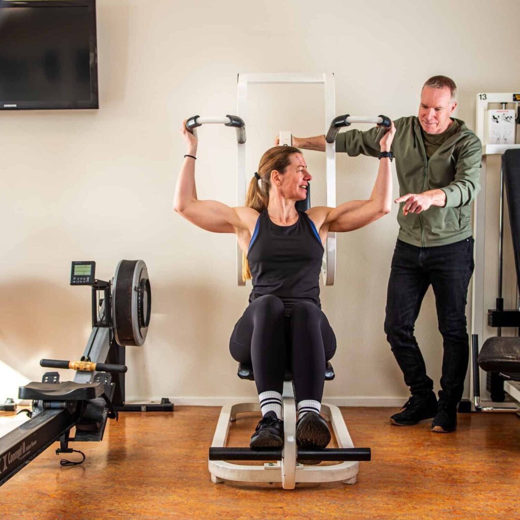 Een vrouw zit op een lat pulldown machine en tilt de hendels op, terwijl een man naast haar staat, glimlacht en instructies geeft in een sportschool.