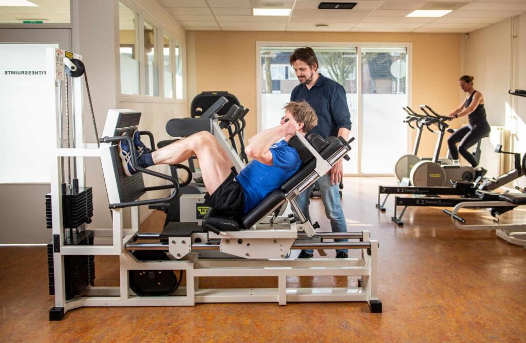 Een man in blauwe sportkleding gebruikt een leg press machine terwijl een andere man toezicht houdt. Op de achtergrond oefent een vrouw op een hometrainer in een lichte sportzaal.