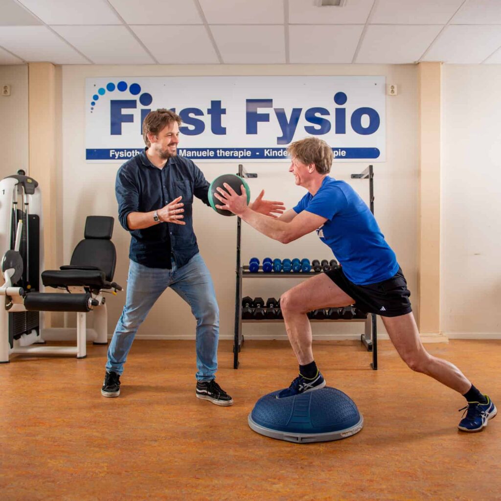 Een man in sportkleding balanceert op een halve gymnastiekbal terwijl hij een medicijnbal vasthoudt. Een andere man in vrijetijdskleding staat voor hem en lijkt hem te coachen of te begeleiden in een fysiotherapiekliniek.