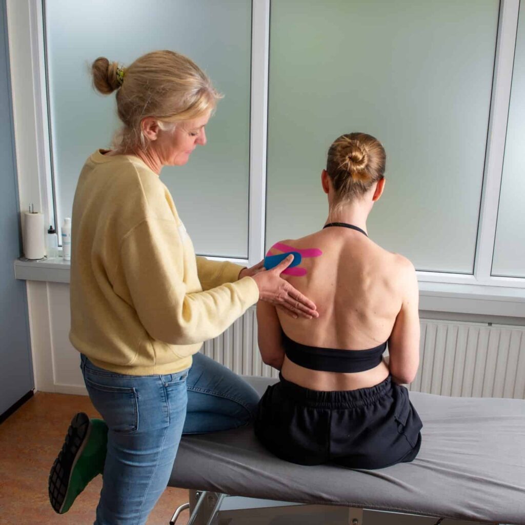 Een vrouw brengt kleurrijke kinesiologie tape aan op de bovenrug van een andere vrouw terwijl ze op een onderzoekstafel zit in een medische of therapeutische ruimte.
