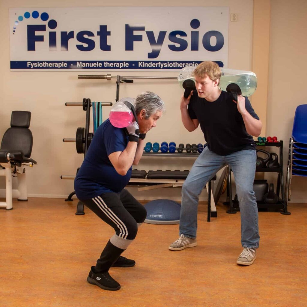 Twee mensen oefenen in een sportschool, ze hurken terwijl ze gewichten op hun schouders houden. Op een bord achter hen staat First Fysio. Fitnessapparatuur en gestapelde stoelen zijn zichtbaar op de achtergrond.