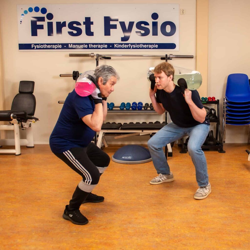Twee mensen voeren binnen squats uit met elk een met water gevuld gewicht op hun schouders. Ze staan tegenover elkaar in een fitness- of fysiotherapiestudio met apparatuur op de achtergrond.
