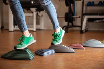 Een persoon met groene gymschoenen stapt op balanspods met textuur die in een rij op de vloer van een sportschool staan, wat evenwichts- of fysiotherapieoefeningen suggereert. Op de achtergrond zijn trainingsapparaten en banken te zien.