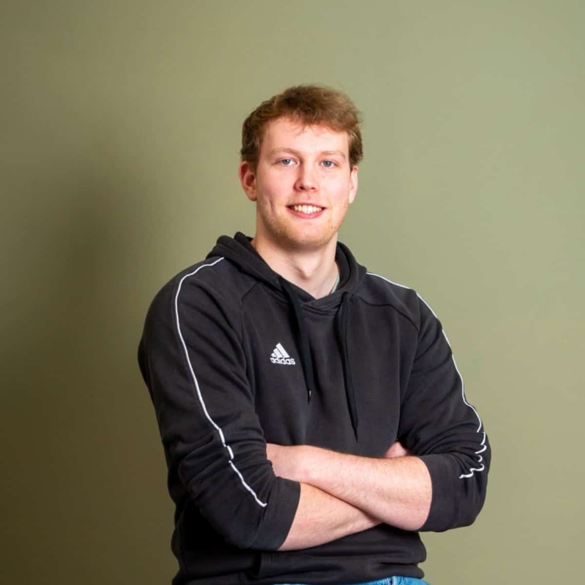 Een jongeman met lichtbruin haar en een zwarte Adidas hoodie zit met zijn armen over elkaar en glimlacht voor een effen olijfgroene achtergrond.