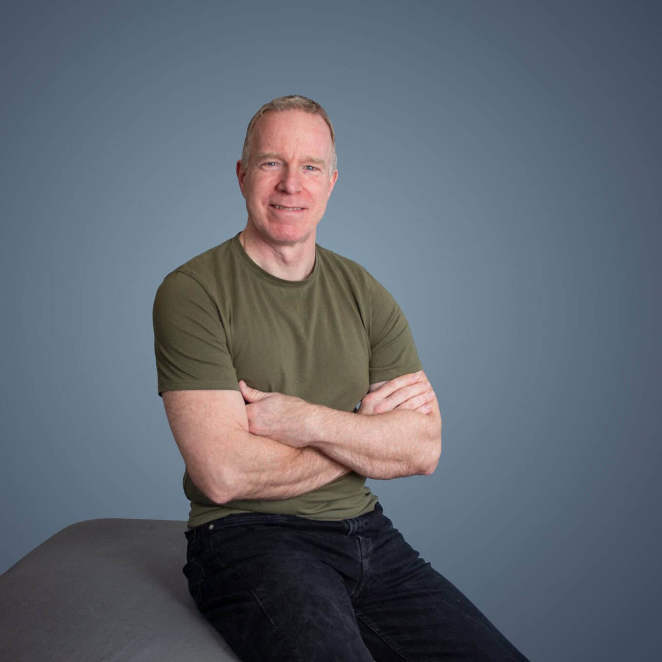 Een man met kort licht haar in een groen t-shirt en een zwarte spijkerbroek zit met zijn armen over elkaar en glimlacht tegen een effen blauwgrijze achtergrond.