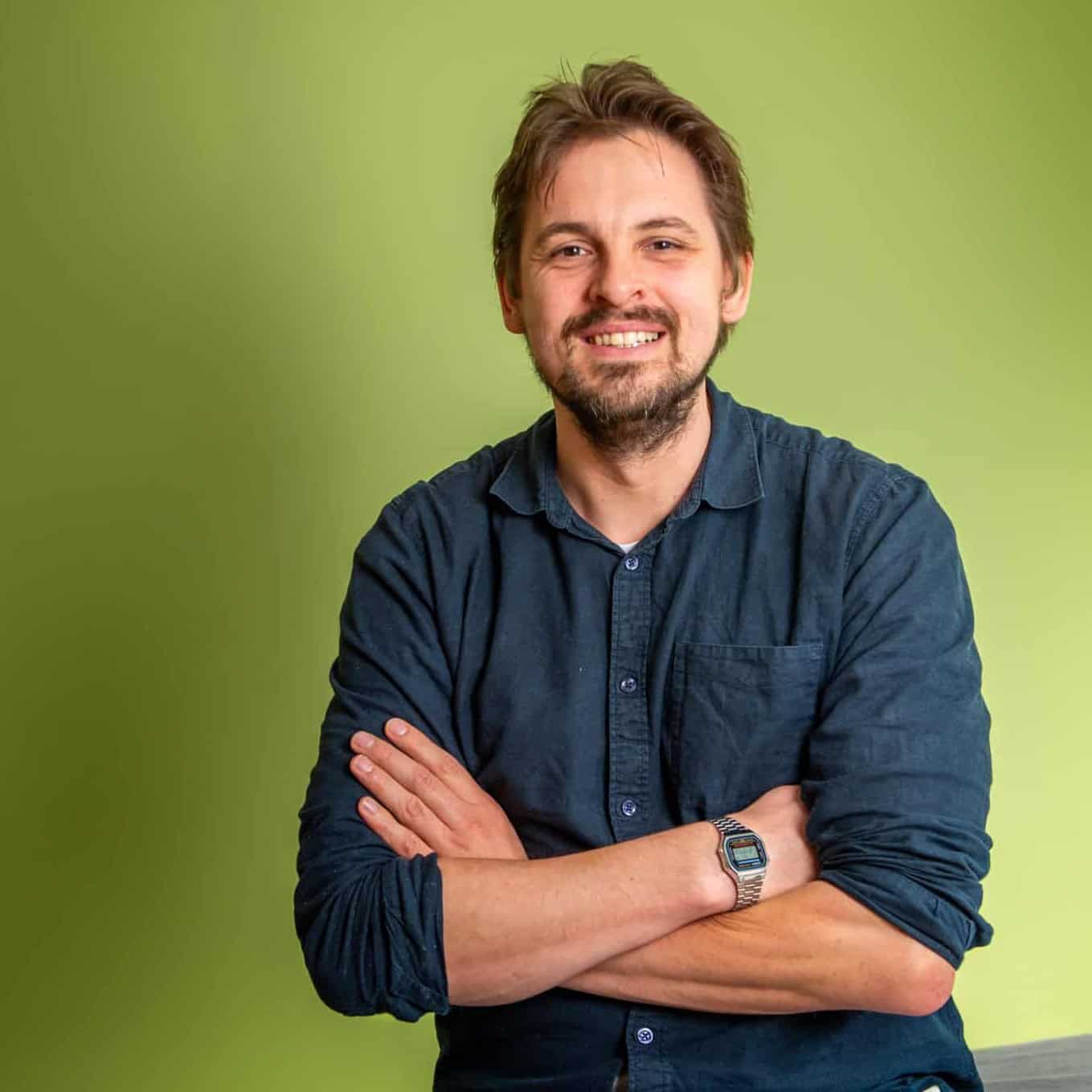 Een man met bruin haar en een baard, een donkerblauw overhemd en een digitaal horloge, glimlacht met zijn armen over elkaar voor een lichtgroene achtergrond.