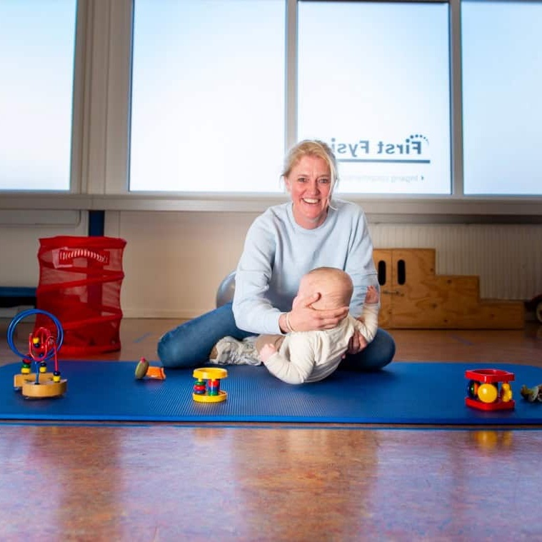 Een glimlachende vrouw zit binnen op een blauwe mat en houdt voorzichtig een baby vast die van zich af kijkt. Kleurrijk speelgoed ligt om hen heen en op de achtergrond zijn grote ramen te zien.